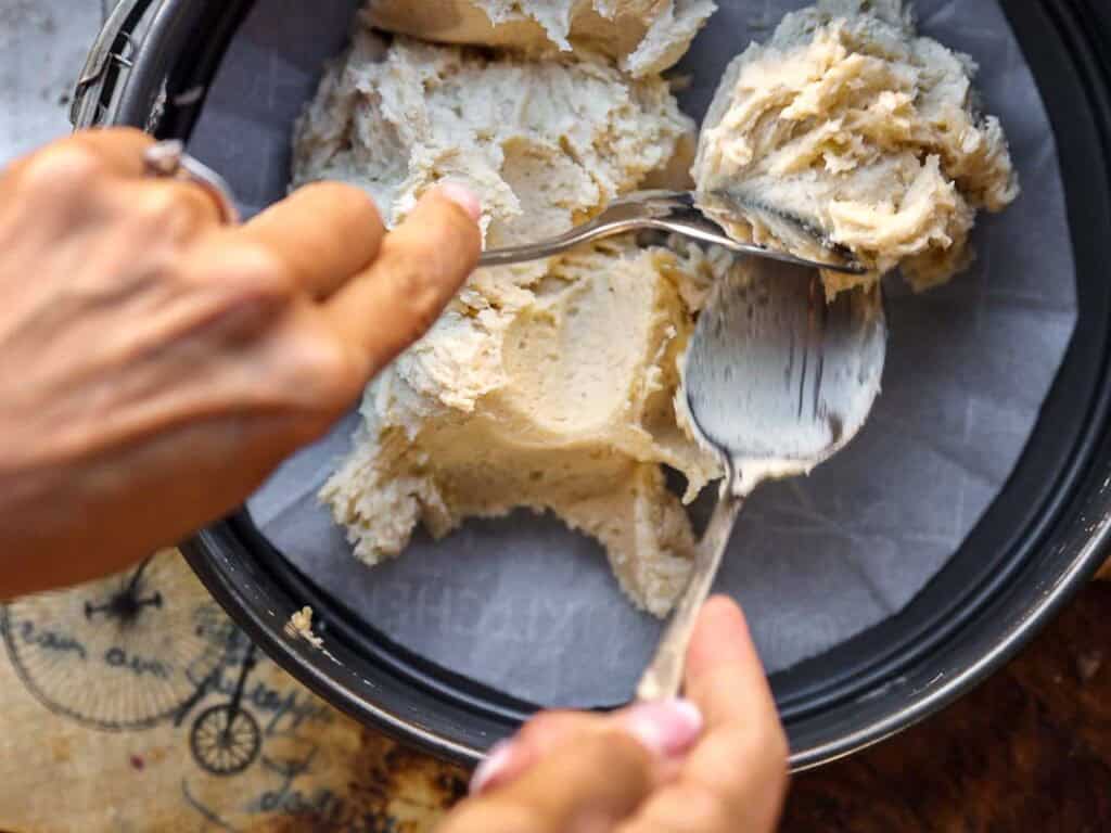 Close-up of hands using two spoons to spread a thick, creamy batter into a round, parchment-lined baking pan. The image suggests preparation for baking, possibly a cake or cheesecake base.
